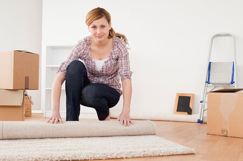 The image depicts a scene in a residential driveway or small yard, where a blue and white Hire Man and Van vehicle is parked on the gravel surface. The van's side panel is partially visible, showing the back door open, revealing a large open cargo area filled with various types of waste, including black rubbish bags, cardboard boxes, and some loose construction debris. In the foreground, there are additional waste items stacked, such as a discarded mattress with a white and light blue cover, a wooden pallet leaning against the van, and some plastic packaging. The surrounding area includes a low brick wall on the left, with a pathway leading towards a garden or backyard space behind the vehicle. The environment appears overcast, with diffuse daylight casting soft shadows and minimal glare. The background hints at a quiet residential neighbourhood, with hints of greenery and neighbouring houses visible, consistent with typical rubbish removal activities handled by Hire Man and Van in the Greenlaw area. The scene emphasizes the practical aspects of waste collection and removal services provided within the local community, with a focus on visible waste materials and the work vehicle ready for debris loading, illustrating an efficient rubbish clearance operation in TD10 postal code surroundings.